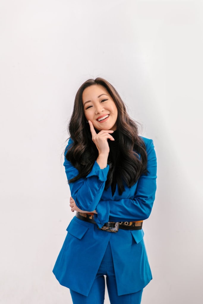 Person with long hair wearing a blue suit smiles with one finger resting on their chin against a plain background.